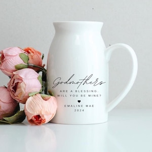 May include: White ceramic pitcher with a curved handle, featuring the text "Godmothers are a blessing. Will you be mine?" and a heart symbol. Pink and peach-colored roses are arranged beside the pitcher.