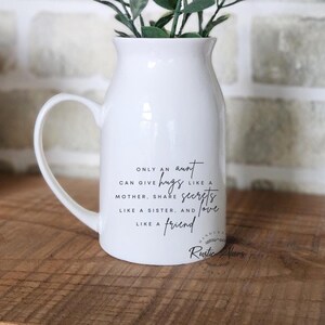 May include: A white ceramic pitcher with a handle, holding greenery. The pitcher has black text that reads "Only an aunt can give hugs like a mother, share secrets like a sister, and love like a friend."