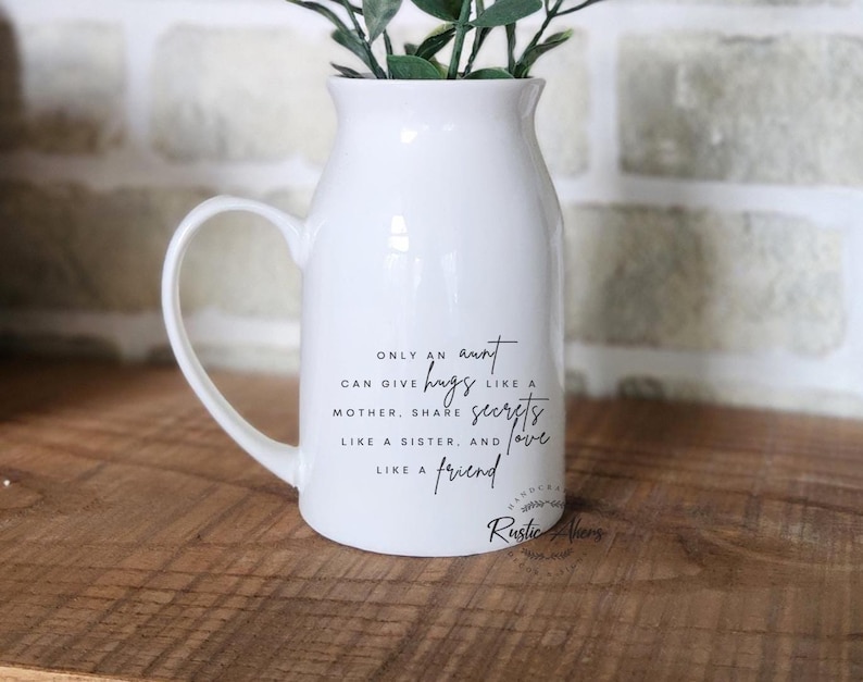 May include: A white ceramic pitcher with a handle, holding greenery. The pitcher has black text that reads "Only an aunt can give hugs like a mother, share secrets like a sister, and love like a friend."