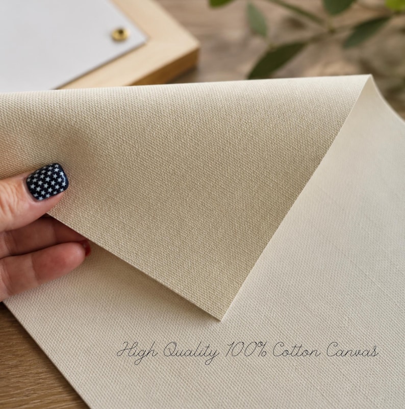 Puede incluir: Una hoja de lienzo de algod&oacute;n texturizado en color blanco roto con un grano tejido visible. La superficie es mate y de color neutro. El texto "High Quality 100% Cotton Canvas" est&aacute; impreso en una fuente cursiva oscura en la parte inferior.
