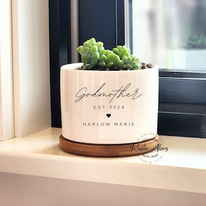 May include: White ceramic planter with the text "Godmother Est 2024" and "Harlow Marie" with a small heart symbol. The planter has a wooden base and is filled with a green succulent plant.