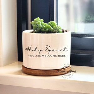 May include: A white ceramic plant pot with the words "Holy Spirit, You Are Welcome Here" in black script. The pot contains a green succulent and rests on a round wooden coaster. The pot is on a white windowsill.