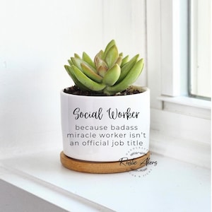 May include: White ceramic succulent planter with a wooden base. The planter has black text that reads "Social Worker because badass miracle worker isn't an official job title". The text "Rustic Akens Decor & Signs" is also visible.