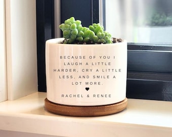 May include: A white ceramic planter with a succulent plant. The planter has the text "BECAUSE OF YOU I LAUGH A LITTLE HARDER, CRY A LITTLE LESS, AND SMILE A LOT MORE. RACHEL & RENEE" and sits on a wooden coaster.