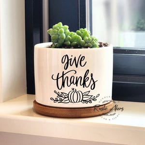Puede incluir: Maceta de cerámica blanca con un diseño en blanco y negro que dice "give thanks" con una calabaza y hojas. La maceta está sentada sobre una base de madera.