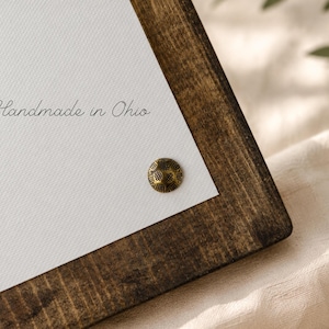 Puede incluir: Un marco de madera marr&oacute;n oscuro que sostiene una superficie de lienzo blanco. El texto "Handmade in Ohio" est&aacute; impreso en cursiva negra sobre el lienzo. Un peque&ntilde;o perno de metal color lat&oacute;n, redondo y texturizado, est&aacute; fijado abajo.
