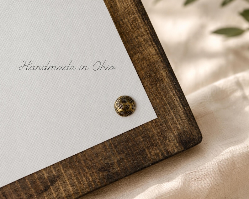 Puede incluir: Un marco de madera marr&oacute;n oscuro que sostiene una superficie de lienzo blanco. El texto "Handmade in Ohio" est&aacute; impreso en cursiva negra sobre el lienzo. Un peque&ntilde;o perno de metal color lat&oacute;n, redondo y texturizado, est&aacute; fijado abajo.