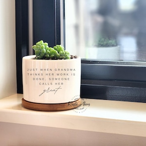 May include: White ceramic planter with a wood base. The planter has a quote that reads "Just when Grandma thinks her work is done, someone calls her great".