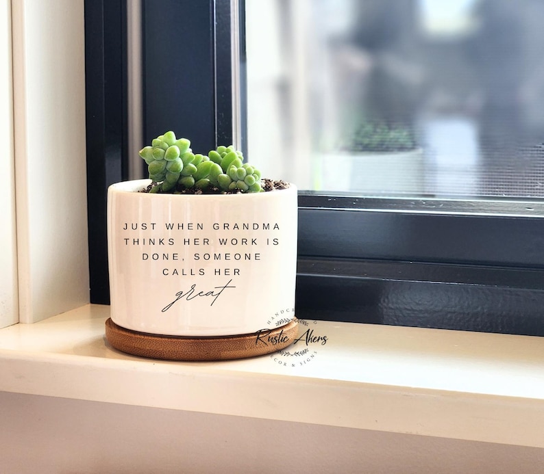 May include: White ceramic planter with a wood base. The planter has a quote that reads "Just when Grandma thinks her work is done, someone calls her great".