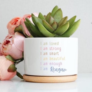 May include: A white ceramic planter with a light wood base containing a green succulent. The planter features the text: I am loved, I am strong, I am smart, I am beautiful, I am enough, I am Reagan, printed in various colors. Pink flowers are beside it.
