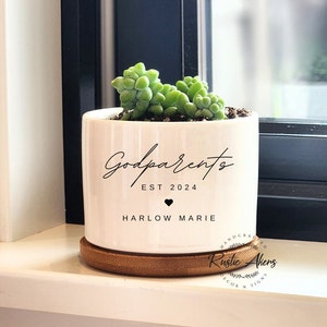 May include: White ceramic planter with the text "Godparents Est 2024 Harlow Marie" and a small succulent plant inside. The planter is sitting on a wooden base.