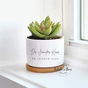 May include: White ceramic planter with a succulent plant. The planter is decorated with the text "Dr. Jennifer Kane Ph.D. 2024" and a small logo that says "Rustic Ahens Decor & Signs".