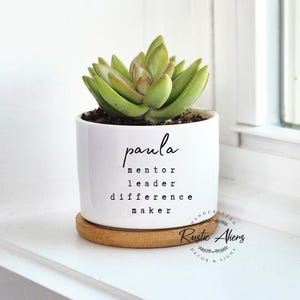 Puede incluir: Maceta de cerámica blanca con una planta suculenta. La maceta tiene el nombre "Paula" escrito en ella, junto con las palabras "mentor, líder, diferencia maker". La maceta está sentada sobre una base de madera y tiene un logotipo de Rustic Ahens en el lateral.