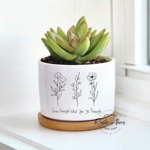 Puede incluir: Una maceta de cerámica blanca con ilustraciones florales negras y el texto "Grow Through What you go Through". La maceta contiene una suculenta verde y descansa sobre un posavasos redondo de madera. Un artículo decorativo.