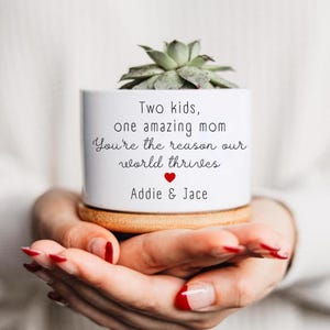 Puede incluir: Una maceta de cerámica blanca con base de madera que contiene una suculenta verde. El texto dice: Two kids, one amazing mom. You're the reason our world thrives. Un pequeño corazón rojo está centrado debajo del texto, seguido de los nombres Addie & Jace.