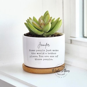 Puede incluir: Una maceta de cerámica blanca con una planta suculenta. La maceta tiene el nombre "Jennifer" y una cita impresa en negro. Se asienta sobre un posavasos redondo de bambú marrón claro. La suculenta tiene hojas verdes y beige.