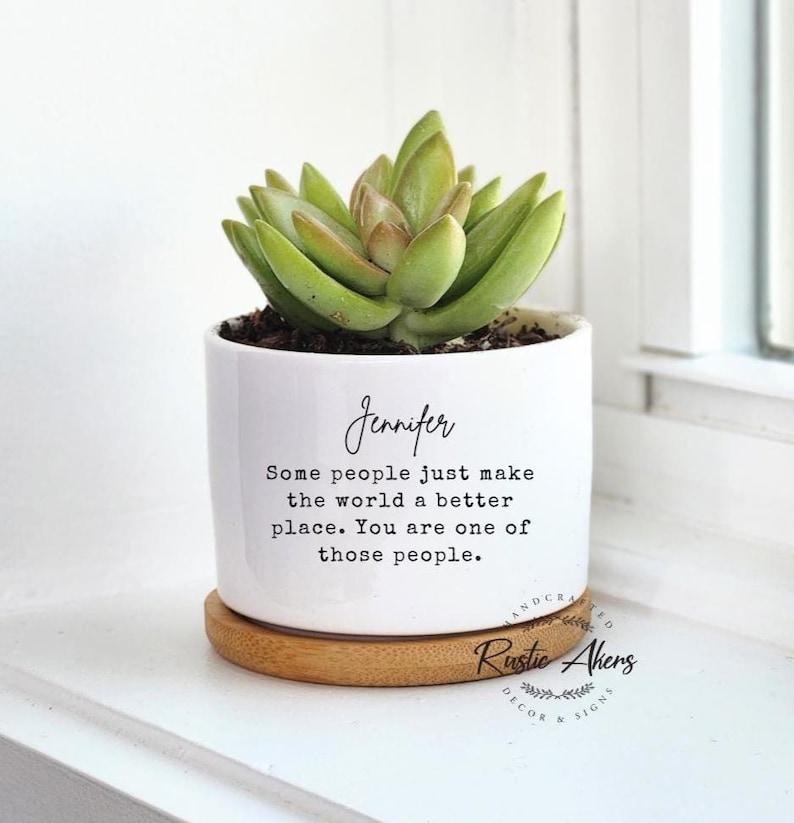 May include: A white ceramic plant pot with a succulent. The pot is inscribed with the name "Jennifer" and a quote: "Some people just make the world a better place. You are one of those people." It rests on a round wooden coaster.