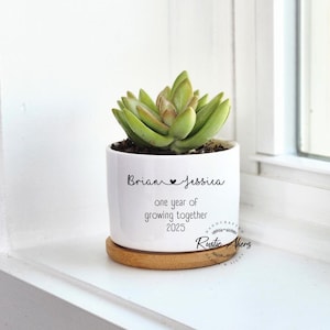 May include: A white ceramic planter with a succulent plant. The planter has the names "Brian & Jessica" and the text "one year of growing together 2025". It sits on a wooden saucer.