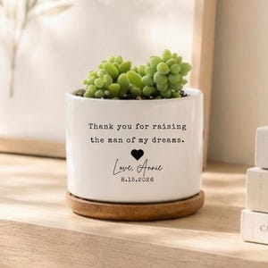 以下が含まれることがあります： 緑色の多肉植物が入った白い円筒形の陶器製プランター。プランターには黒い文字で「Thank you for raising the man of my dreams. Love, Annie 8.15.2026」と印刷されており、名前の上には小さな黒いハートのアイコンが描かれています。