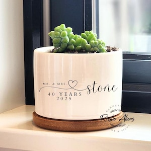 Peut inclure: Pot en céramique blanche avec une base en bambou. Le pot a un texte noir qui dit "Mr. & Mrs. stone 40 Years 2025". Le texte est en écriture cursive avec un symbole de cœur. Le pot est rempli d'une petite plante succulente.