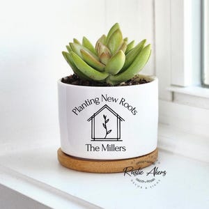 May include: A white ceramic planter with a succulent plant. The planter has the text "Planting New Roots" and "The Millers" in black. It sits on a round, light brown wooden coaster. The succulent has green and tan leaves.