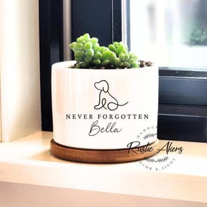 May include: White ceramic planter with a succulent plant. The planter has a black line drawing of a dog and the words "NEVER FORGOTTEN" and "Bella". The planter sits on a round, wooden coaster. The words "Rustic Akers" are also visible.