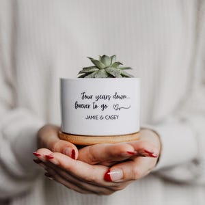 May include: A white ceramic planter with a succulent plant, on a wooden coaster. The planter has the text "Four years down... forever to go" and the names "JAMIE & CASEY" in black script. The planter is held in open hands.