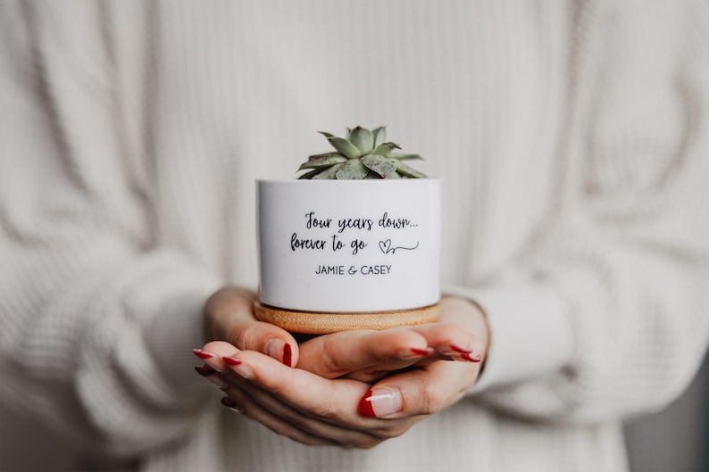 May include: A white ceramic planter with a succulent plant, on a wooden coaster. The planter has the text "Four years down... forever to go" and the names "JAMIE & CASEY" in black script. The planter is held in open hands.