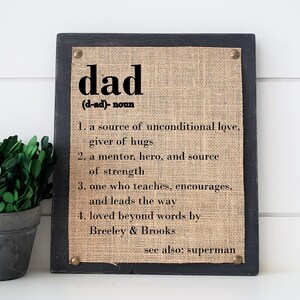 May include: A framed burlap sign with the word "dad" defined as a source of unconditional love, giver of hugs, mentor, hero, and source of strength. The sign also says "one who teaches, encourages, and leads the way" and "loved beyond words by Breeley & Brooks". The sign also says "see also: superman".