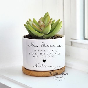 May include: White ceramic planter with a succulent plant. The planter has the text "Mrs. Stevens Thank you for helping me grow. Madison" with a black heart.