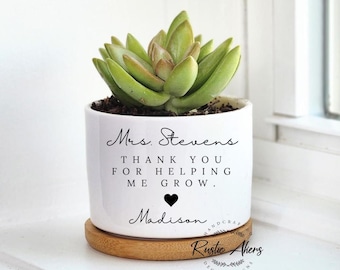 May include: A white ceramic planter with a succulent plant. The planter has the text "Mrs. Stevens, THANK YOU FOR HELPING ME GROW, Madison" in black script. It sits on a round wooden saucer. The words "Rustic Akens, DECOR & SIGNS" are also visible.
