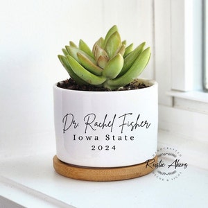 May include: White ceramic succulent planter with the text "Dr. Rachel Fisher Iowa State 2024" and a wooden base. The planter is decorated with a green succulent plant. The text "Handcrafted Rustic Ahens Decor & Signs" is printed on the base.