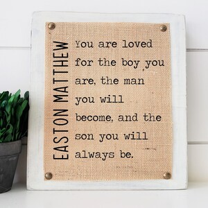 May include: A white wooden sign with a burlap background and the text "EASTON MATTHEW You are loved for the boy you are, the man you will become, and the son you will always be."