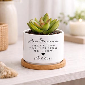 May include: A white ceramic planter with a succulent plant, sitting on a round wooden coaster. The planter has the text "Mrs. Stevens, THANK YOU FOR HELPING ME GROW" with a heart and the name "Maddie".
