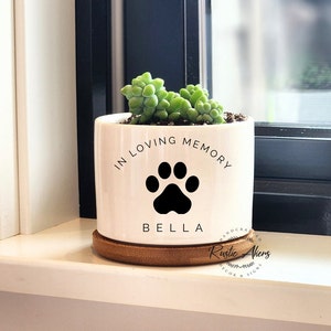 May include: White ceramic planter with the text "In Loving Memory" and a black paw print. The name "Bella" is written below the paw print. The planter is sitting on a wooden base.