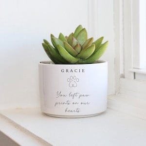 May include: White ceramic planter with the name "GRACIE" and a paw print. The text reads "You left paw prints on our hearts". A green succulent plant is in the planter.