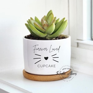 May include: A white ceramic planter with a succulent plant. The planter has a cat face design with the words "Forever Loved" and "Cupcake". The planter sits on a round, wooden saucer.