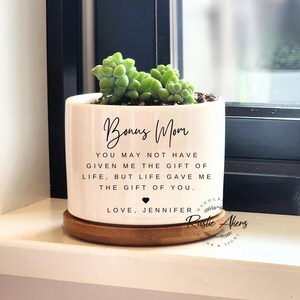 以下が含まれることがあります： 白いセラミック製のプランターに多肉植物と「Bonus Mom You may not have given me the gift of life, but life gave me the gift of you. Love, Jennifer」というテキストが書かれています