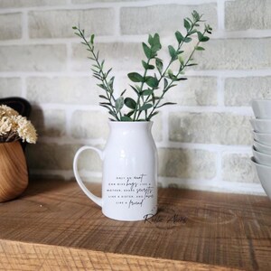 May include: A white ceramic vase with a handle, filled with green leafy sprigs. The vase has a quote that reads "Only an aunt can give hugs like a mother, share secrets like a sister, and love like a friend." The vase is on a wooden shelf.