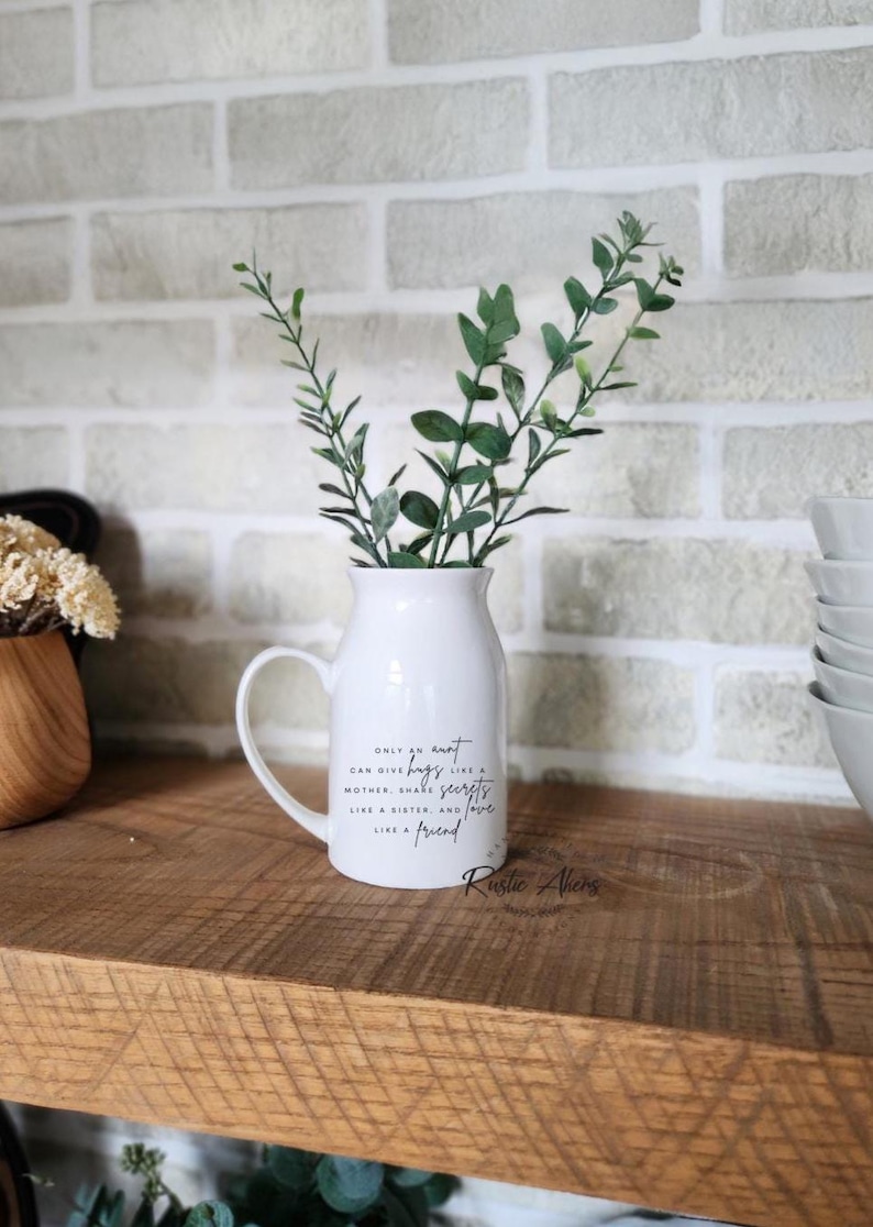 May include: A white ceramic vase with a handle, filled with green leafy sprigs. The vase has a quote that reads "Only an aunt can give hugs like a mother, share secrets like a sister, and love like a friend." The vase is on a wooden shelf.