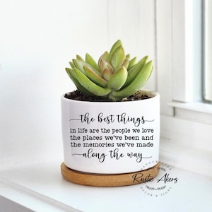 May include: White ceramic planter with a succulent plant and the text "the best things in life are the people we love the places we've been and the memories we've made along the way". The planter has a wooden base and the text "HANDCRAFTED Rustic Akers DECOR & SIGNS" is printed on the base.