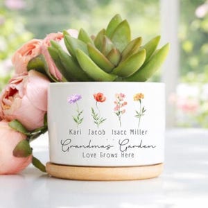 May include: A white ceramic planter with a succulent plant, featuring floral illustrations and the text "Grandma's Garden Love Grows Here." The planter sits on a wooden saucer, with pink roses in the background.