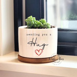 May include: White ceramic planter with a succulent plant and the text "sending you a hug" with a red heart. The planter has a wooden base.
