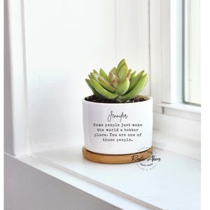 May include: A white ceramic plant pot with a succulent, on a small wooden coaster. The pot has the name "Jennifer" and a quote in black script. The succulent is green with a touch of brown.
