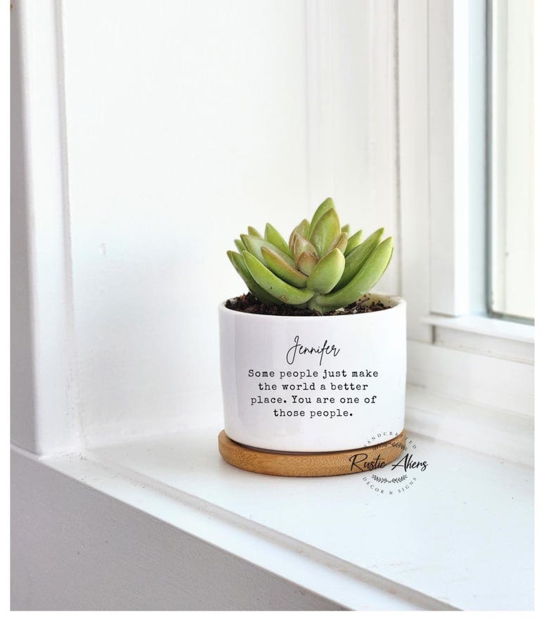 May include: A white ceramic plant pot with a succulent, on a small wooden coaster. The pot has the name "Jennifer" and a quote in black script. The succulent is green with a touch of brown.