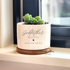 May include: White ceramic planter with a wooden base. The planter has the text "Godfather EST 2024" and "HARLOW MARIE" with a heart symbol in between. A small green succulent plant is in the planter.