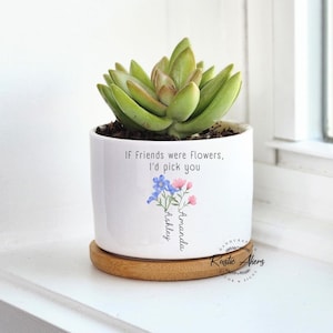May include: A white ceramic planter with a succulent plant. The planter has a floral design with the text "If friends were flowers, I'd pick you" and the names "Ashley" and "Amanda". The planter sits on a round wooden coaster.