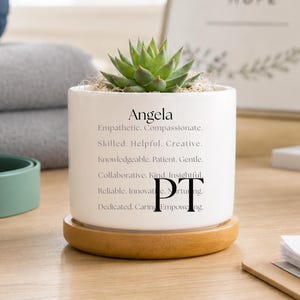 May include: A white ceramic cylindrical planter with a light wood saucer base containing a green succulent. The planter features the name Angela followed by a list of descriptive words and the large black letters PT printed on the front surface.