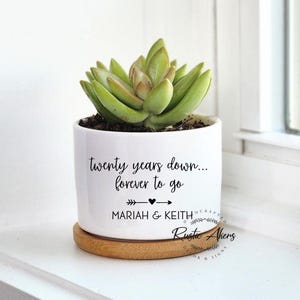 May include: A white ceramic planter with a succulent plant. The planter has the text "twenty years down... forever to go" with the names "MARIAH & KEITH" below. It sits on a round wooden coaster.
