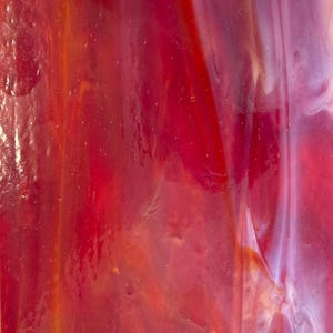 May include: Abstract close-up of stained glass with a blend of red, orange, and white hues. The glass has a glossy finish, with streaks and variations in color creating a dynamic visual texture. The light plays across the surface, highlighting the color variations.